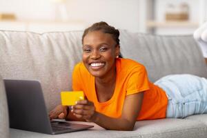 Joyful Afro woman shopping online with laptop pc and credit card, lying on couch at home. Millennial black lady searching web for goods on dicsount, buying at internet store, ordering remotely photo