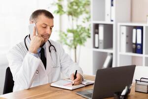 Making an Appointment With a Doctor Concept. Clinician talking on phone with his patients and writing in notebook photo