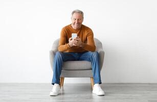 Front portrait of smiling mature man using mobile phone, watching sitting on armchair isolated on white studio background wall. Guy browsing internet, surfing web, free copy space photo