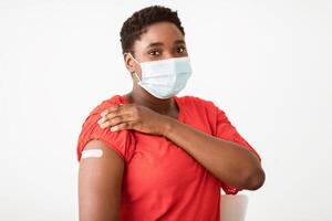 I'm Vaccinated. Black Woman Showing Arm With Plaster Strip After Getting Covid-19 Coronavirus Vaccine Injection Posing On White Studio Background, Wearing Protective Face Mask. Covid Vaccination photo