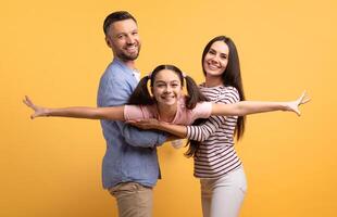A father, mother, and daughter share a joyful moment with the girl extending her arms as if flying. Their expressions reflect love and affection, showcasing a close family bond. photo
