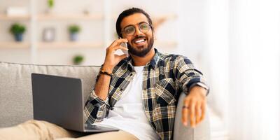 Joyful middle-eastern young man freelancer working from home, sitting on couch with laptop, having phone conversation with clients or colleagues, smiling and looking at copy space, remote job concept photo