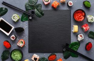 Top view on traditional japanese empty square black slate plate framed with sushi, ginger, soy, wasabi bowls, green leaves and chopsticks on rustic grey backround copy space photo