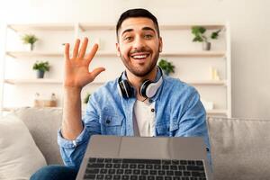 Online Call. Portrait of smiling bearded man having virtual conference with friends, family or colleagues using computer, waving hello with hand, sitting on couch at home office, webcamera view photo