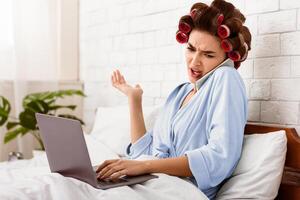 Discontented Housewife Talking On Cellphone Using Laptop Computer Lying In Bed At Home. Displeased Customer Concept photo