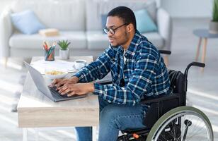 Online work for disabled people. Handicapped black guy in wheelchair using laptop for remote job at home. Millennial paraplegic man having business meeting on internet, making call photo
