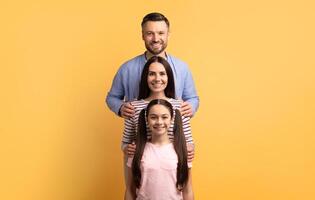 A family of three is standing together against a bright yellow background. The father, mother, and daughter show love and affection, smiling happily and bonding in this joyful moment. photo