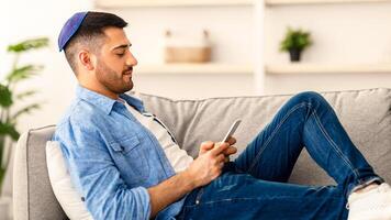 Rest With Gadgets. Side view profile of millennial jewish guy in yarmulke using mobile phone at home, messaging or browsing social media, sitting on couch in living room, checking email photo