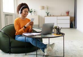 People And Technology. Smiling young black lady sitting at tea table with laptop, holding using cell phone. Cheerful female in wireless headset watching , chatting taking break, free copy space photo