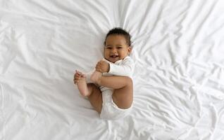 Cute Funny Little Black Baby Having Fun While Lying On Bed At Home, Adorable African American Infant Boy Playing With His Toes And Laughing While Resting In Bedroom, Top View, Copy Space photo
