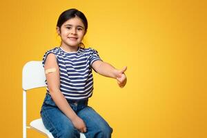 Kids Vaccination. Vaccinated Little Arab Girl Gesturing Thumb Up After Covid-19 Vaccine Injection, Cheerful Middle Eastern Child With Adhesive Band On Arm Sitting On Chair Over Yellow Background photo