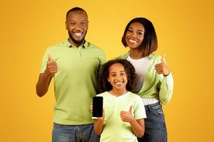 Positive african american family of three demonstrating smartphone and showing thumbs up, posing over yellow background. Happy parents and child recommending new mobile application or website photo