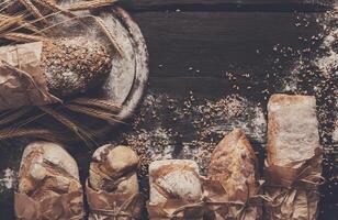 Bread background. Brown and white whole grain loaves wrapped in kraft paper composition on rustic dark wood with wheat ears scattered around. Baking and home bread making concept. Soft toning photo