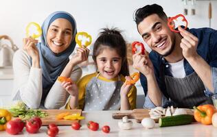 Portrait Of Happy Arab Family With Little Daughter Holding Bell Pepper Slices While Posing In Kitchen Together, Cheerful Middle Eastern Family Of Three Having Fun While Coocking Healthy Food At Home photo