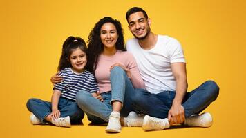 Portrait Of Happy Young Middle Eastern Family Of Three With Little Daughter Posing Together In Studio, Cheerful Arab Parents And Cute Child Sitting On Floor And Smiling At Camera, Free Space photo