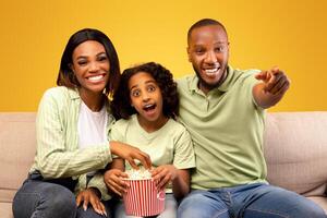 Excited black parents and their little daughter eating popcorn and watching movie on TV, man pointing at camera, sitting on couch over yellow studio background photo