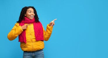 African American Lady In Winter Clothes Pointing Fingers Aside Advertising Text Standing Over Blue Studio Background. Look There, Advertisement Banner. Panorama With Empty Space For Text photo