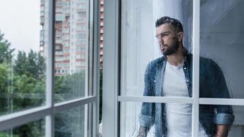 Depressed sad man looking through window, hoping for end of pandemic. Sad unhappy millennial european handsome guy with crossed arms looking at large window with drops in kitchen interior, empty space photo