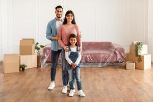 House Move, Renovation And Repair Concept. Full body length of cheerful young family of three people standing in new empty apartment, posing and looking at camera, hugging, free copy space photo