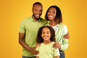 Family love concept. Portrait of black family with daughter hugging and smiling at camera standing isolated on yellow background. Mum and dad posing with child in studio photo