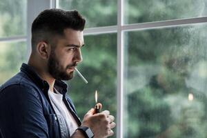 Very depressed man destroyed, sad, suffering emotional crisis and grief thinking of difficult and hard decision. Unhappy young european attractive guy lighting cigarette, on large window background photo