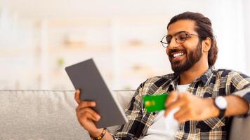 Closeup of handsome smiling arab guy banking online from home while lockdown, chilling on couch, holding credit card and modern digital tablet, using financial mobile application, copy space photo