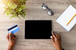 Top view of Afro lady with credit card using tablet pc for online shopping at wooden table, mockup for website or app. Millennial black woman ordering goods on touch pad, making contactless payment photo