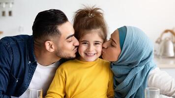 Loving Parents Kissing Their Little Daughter To Both Cheeks While Eating Breakfast In Kitchen, Happy Middle Eastern Dad And Mom In Hijab Bonding With Cute Female Child At Home, Enjoying Time Together photo