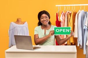 Smiling young black lady sitting at table with laptop, showing OPEN sign, posing near clothing rack on orange studio background. Concept of online shopping for summer clothes photo