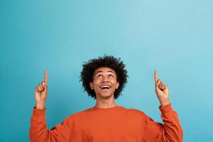 A young African American man with curly hair wears an orange sweater and smiles widely while pointing upward at copy space with both hands. The vibrant blue background enhances his joyful expression. photo