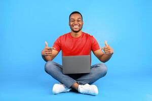 Successful Black Male Freelancer Pointing Fingers At Laptop Smiling To Camera Sitting Over Blue Background. Studio Shot Of Happy Millennial Guy Posing With Computer. Technology Concept photo