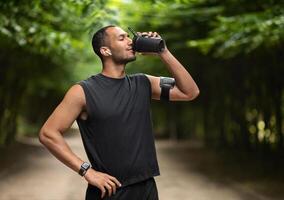 Sporty young black man drinking protein cocktail, training at public park, using wireless earbuds, listening to music while jogging by forest, copy space. Sports nutrition concept photo