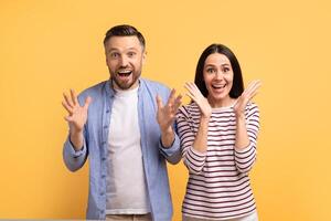 A Caucasian couple stands together, expressing excitement and joy with wide smiles and open hands. The bright yellow background enhances their affectionate bond, capturing pure happiness. photo