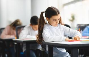 Portrait of small girl with ponytails sitting at table in classroom at primary school or kindergarten, writing or drawing in notebook. Reopening and return back to school after coronavirus quarantine photo