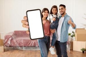 Moving Day. Portrait of cheerful daddy, mum and daughter posing in new apartment, standing in empty living room showing thumbs up sign gesture. Cheerful parents holding girl on hands, free copy space photo