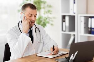 General Practitioner Concept. Portrait of middle aged doctor physician talking on mobile phone with his patient and writing in his diary making notes or appointment, consulting client. Telemedicine photo