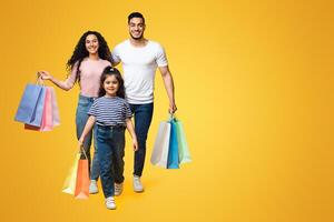 Shopping Day. Cheerful Young Middle Eastern Family Of Three Walking With Bright Shopper Bags Over Yellow Background In Studio, Happy Arab Parents And Female Child Enjoying Seasonal Sales, Copy Space photo