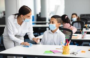 Hygiene And Cleaning. Teacher cleaning table with antibacterial sanitizer and napkin, disinfecting surface, applying antiseptic spray on desk, wearing disposable face mask, boy studying in classroom photo