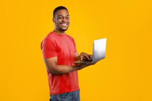 Freelance Career. Happy Black Guy Using Laptop Computer Smiling To Camera Standing Over Yellow Background, Studio Shot. People And Gadgets, E-Learning And Internet Technologies Concept photo