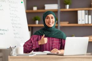 Online Teacher, Distant Learning, Recording Vlog. Smiling Middle Eastern lady tutor in headscarf showing thumbs up gesture, posing looking at camera, sitting at desk at home photo