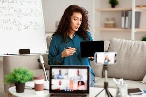 E-Learning. Confident woman having conference call using laptop. Lady teacher showing digital tablet, recording tutorial with mobile phone on tripod, streaming live to diverse students photo