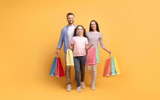 A happy family consisting of a father, mother, and daughter stands together holding colorful shopping bags against a cheerful yellow backdrop. They share smiles and affection. photo
