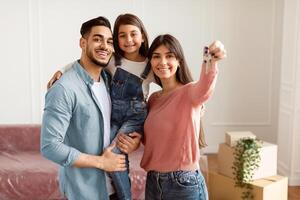 Portrait Of Happy Young Family Showing Key, Cheerful Parents Hugging Little Kid, Holding Daughter On Hands. Smiling People Moving In New Apartment Standing In Living Room. Selective Focus photo