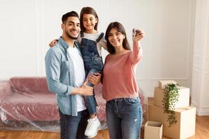 House Ownership. Portrait Of Happy Middle Eastern Family Showing Key, Standing In Living Room. Cheerful Dad Holding Daughter On Hands, People Moving In New Apartment, Posing, Blurred Background photo