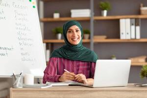 Remote Study Concept. Portrait of smiling arabian woman in hijab sitting at desk with pc laptop near whiteboard. Happy lady posing and looking at camera, writing, taking notes in paper notebook photo