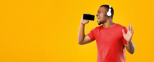 Black Man Singing Holding Smartphone Like Microphone Using Karaoke App Having Fun Standing On Yellow Studio Background. Guy Wearing Wireless Headphones Enjoying Music Online Using Application photo