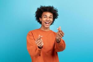 A cheerful African American man showcases a playful pose with hand gestures that symbolize affection. He wears an orange sweatshirt against a vibrant blue backdrop, exuding positivity. photo