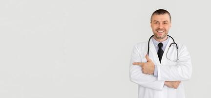 Useful Information. Portrait of smiling confident mature general practitioner pointing away at copy space standing isolated over white studio background, looking at camera. Doctor wearing stethoscope photo