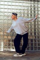 A young man in a gray sweatshirt and black pants dances in front of a glass block wall photo