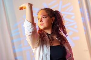 A young woman with curly hair and a white shirt poses with her arm raised in a studio setting, lit by colorful lights photo